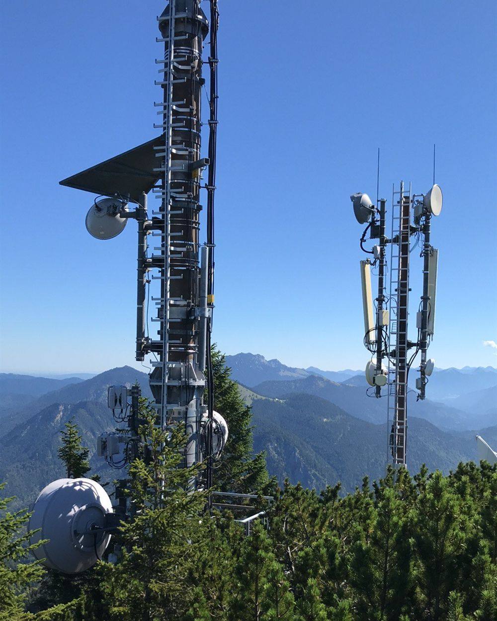 Freigeschnittener Sendemast auf einem Berggipfel, davor Latschenkiefern und die Bergkette