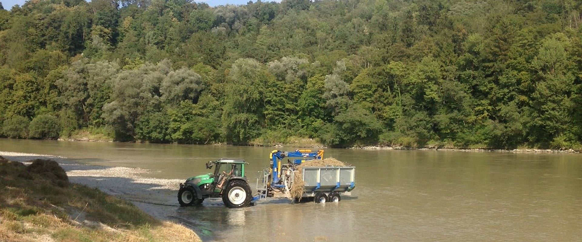 Traktor mit Ladekran bei der Uferpflege im Flussbett, dahinter bewaldete Hänge