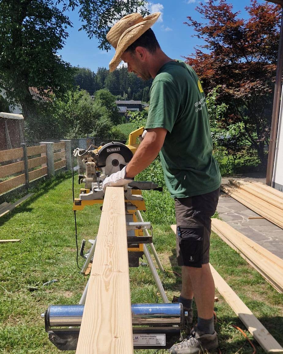 Mitarbeiter mit Strohhut schneidet an der Kappsäge einen langen Holzbalken auf Länge