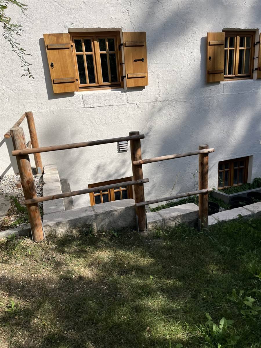 Natursteinmauer und Geländer aus entrindetem Rundholz vor einem weiß verputzten Altbau